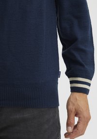 Sötétkék pulóver puha anyagból, bordázott aljjal és mandzsettákkal, krémcsíkokkal. Címke látható az oldalon.