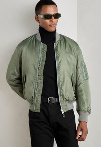 Giacca bomber verde oliva, materiale in nylon morbido, polsini a coste, tasca laterale con zip, abbinata a un dolcevita nero e pantaloni neri.