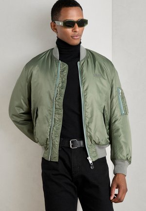 Olijfkleurige bomberjack, smooth nylon materiaal, ribgebreide manchetten, ritszak op de zij, gecombineerd met een zwarte coltrui en zwarte broek.