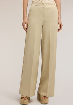 Pantalon beige à jambes larges porté avec des sandales compensées beiges, montré sur une personne debout sur un fond neutre.