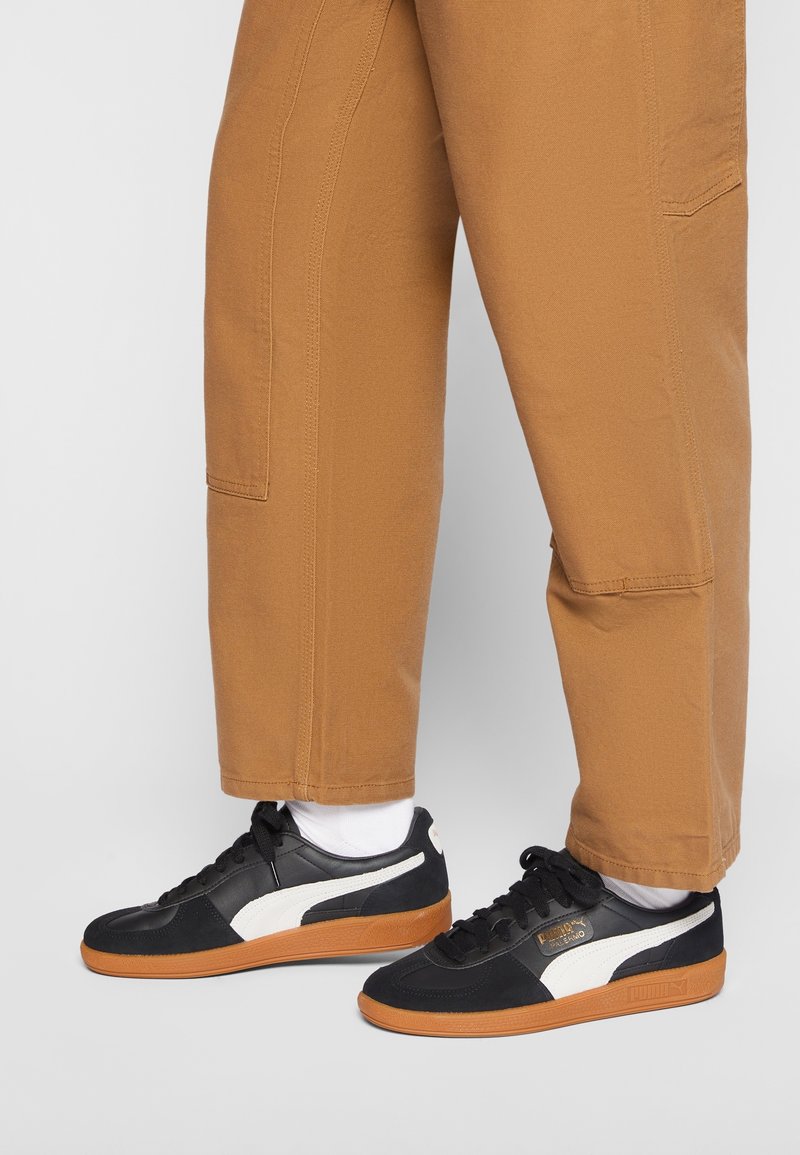 Baskets noires avec des accents blancs et des semelles en caoutchouc naturel, associées à un pantalon ample beige. Elles présentent un design à lacets et un détail de logo sur le talon.