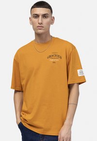 T-shirt en coton jaune moutarde à manches courtes, avec du texte imprimé et une étiquette sur la manche gauche. Coupe droite et texture douce.