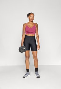 Rosa idrettsbh med "UNDER ARMOUR" logo, svarte høyt liv shorts, holder en svart manual, iført grå treningssko og svarte sokker.
