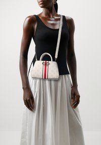 Sac à main blanc avec une rayure rouge, présentant une forme structurée, un accent de logo et une sangle amovible. Le modèle porte un haut noir et une jupe blanche.