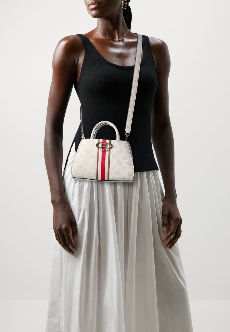 Sac à main blanc avec une rayure rouge, présentant une forme structurée, un accent de logo et une sangle amovible. Le modèle porte un haut noir et une jupe blanche.