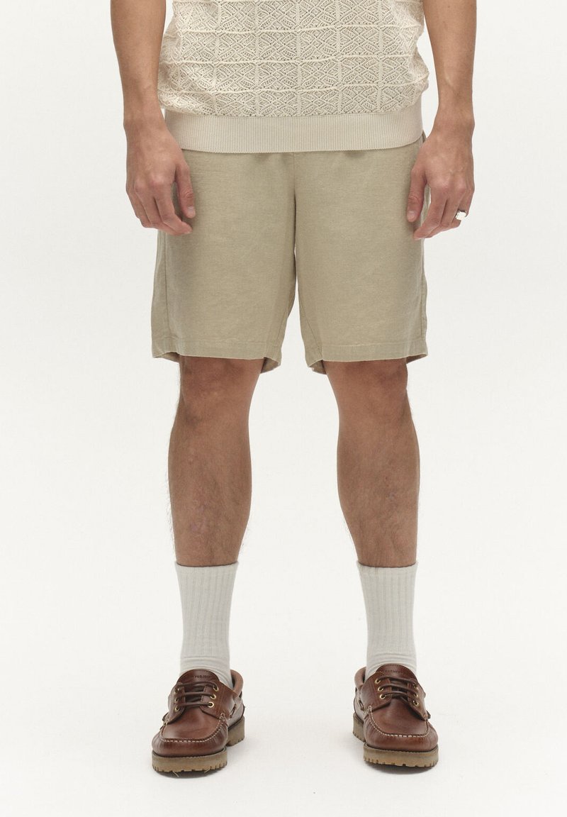 Homme portant un short en maille beige, des chaussettes blanches côtelées, des chaussures bateau en cuir marron, et un pull en maille crème, debout devant un fond uni.