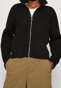 Sudadera negra de punto con cremallera y textura acanalada, y cuello alto. Cuenta con una cremallera plateada y un corte relajado. Combinada con pantalones beige.