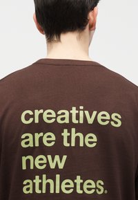 Persona con cabello corto y oscuro que lleva una camisa marrón con texto verde pálido en la espalda: "los creativos son los nuevos atletas".