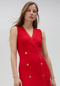Rode mouwloze jurk met een geribde structuur, V-halslijn en decoratieve gouden knopen langs de taille en rok. Soepele stof.