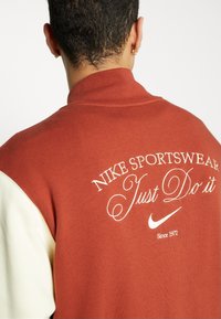Sweatshirt bordeaux avec des accents crème, portant le texte brodé blanc "NIKE SPORTSWEAR Just Do It" et un petit logo Nike, design de col traditionnel.