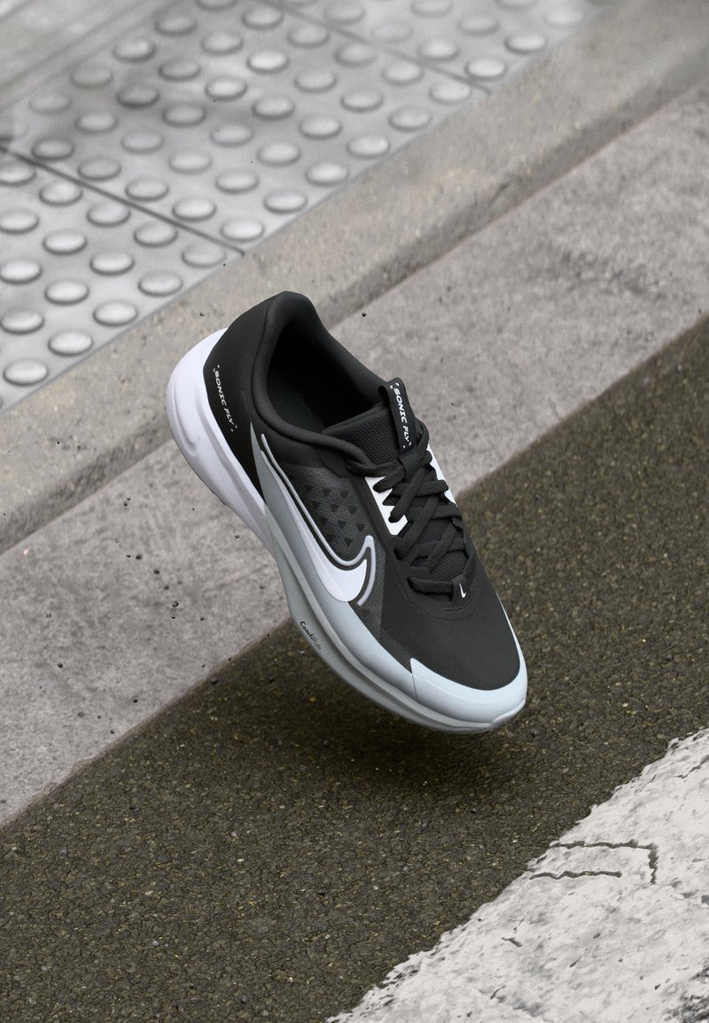 Fekete sportcipő, texturált felsőrésszel, fehér részletekkel és párnázott talppal. A dizájn tartalmaz egy logót és könnyű felépítést.