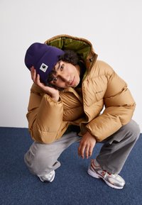 Puffjakke i tan med grøn indvendig, ribbede grå bukser, lilla hue med logo og sølvfarvede sneakers med orange detaljer.