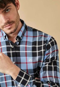 Camisa de cuadros abotonada en azul, negro y rojo. Presenta un cuello, mangas largas y un parche con el logo. La tela parece texturizada con un patrón de cuadros.