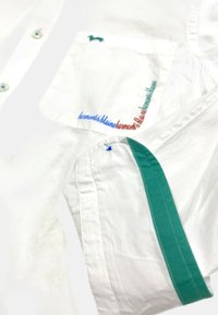 Witte shirt met een groene bies, blauwe en rode tekst op de zak en een klein groen logo. Gemaakt van katoen met een soepele textuur.