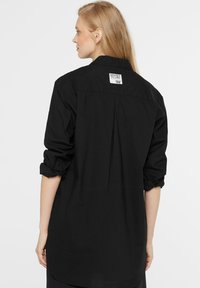 Black shirt with a relaxed fit, rolled sleeves, and a back pleat. Features a fabric label at the neck. Smooth texture, casual design.