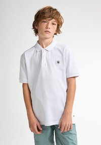 Chemise polo blanche en coton avec un col classique et des manches courtes. Présente un petit logo brodé sur la poitrine. Conception simple, texture lisse.