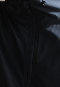 Gros plan d'une veste noire avec une fermeture éclair verticale et une petite bascule sur le cordon près du col.