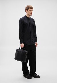 Fekete bőr aktatáska dupla fogantyúval, egy fekete, testhez simuló, gombos ingben és laza fekete nadrágban lévő férfi modellen viselve. Sima textúra.