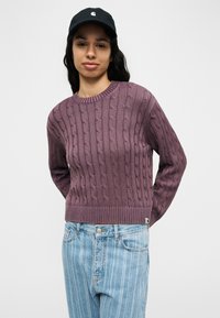 Νεαρή γυναίκα που φοράει μαύρο καπέλο, μοβ πουλόβερ πλεκτό με κοτσίδες και ανοιχτό μπλε τζιν με ρίγες, στέκεται μπροστά σε λευκό φόντο.