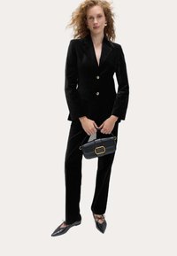Czarna aksamitna marynarka z złotymi guzikami, pasujące spodnie i czarne baleriny. Modelka trzyma małą czarną torebkę z złotymi akcentami.