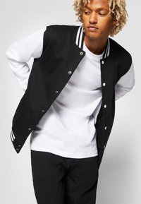 Chaqueta varsity negra con mangas blancas, cuello rayado, botones de presión y bolsillos visibles, llevada sobre una camiseta blanca y pantalones negros.