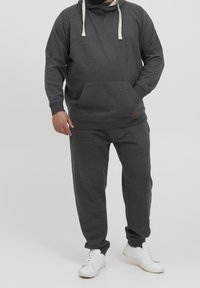 Sudadera gris con capucha ajustable, bolsillo frontal y pantalones de chándal a juego. La tela parece suave y con un corte relajado. Zapatillas blancas completan el conjunto.