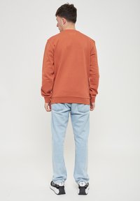 Felpa color ruggine con polsini e orlo a coste, indossata sopra jeans azzurri chiari. Il retro presenta un logo goffrato discreto all'altezza del collo.