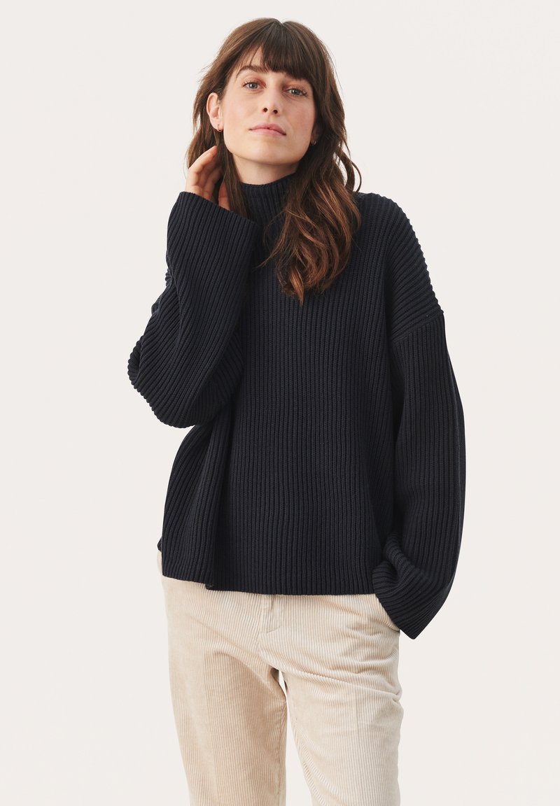 Pulover navy cu dungi, cu gât înalt și mâneci largi, realizat dintr-un material tricotat texturat, combinat cu pantaloni din catifea bej deschis.