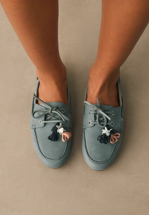 Picioare purtând pantofi de barcă bleu deschis cu șireturi și ciucuri decorative negre, albe și piersică pe un podea neutră.