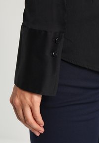 Poignet de chemise noir avec deux boutons, texture lisse et ourlet soigné. Main positionnée sur un tissu marine, mettant en valeur les détails du poignet.