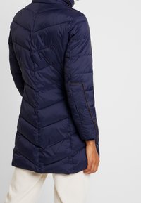 Námořnicky modrá péřová bunda s cikcak prošitím, kapsou na zip po straně a kontrastní hnědou lemovkou na rukávech a kapsách. Střední délka.