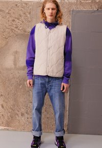 Gepolsterte hellgraue Weste über einem lila Hoodie, kombiniert mit gerollten Jeans und schwarzen Turnschuhen mit lila Akzenten. Strukturierter und geschichteter Look.