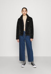 Giacca in pelliccia sintetica nera con collo a camicia, abbinata a un dolcevita beige e jeans a gamba larga blu. Sneakers nere alte.
