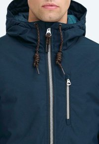 Uomo che indossa una giacca con cappuccio blu navy, dotata di lacci neri e arancioni, zip anteriore argentata e tasca sul petto con zip verticale.