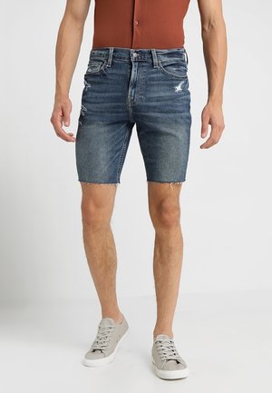 Donkerblauwe jeansshorts met een versleten ontwerp, rafelige zoom en klassieke vijf-pocketstijl. Gepaard met lichtgrijze sneakers.