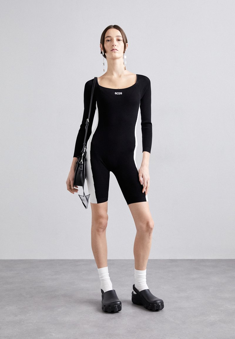 Black bodysuit with long sleeves, square neckline, white side panels; paired with black clogs and white crew socks, holding a black bag.