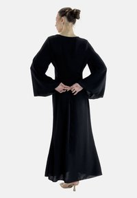 Langes schwarzes Kleid mit fließenden, Glockenärmeln. Glatter Stoff, taillierte Passform und ein ausgestellter Rock. Eine Rückenschnürung verleiht dezente Eleganz.