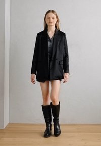 Crni dvoredni sako od glatke tkanine, uparen s tamnom košuljom i šortsevima. Čizmice od crne kože do koljena upotpunjuju izgled.