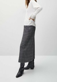 Jupe à carreaux gris avec une silhouette longue en A. Associée à un pull blanc, portée avec des bottines noires. Détails en tissu texturé visibles.