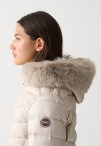 Giacca imbottita beige con cappuccio foderato in pelliccia, design trapuntato e toppa con logo tondeggiante sulla manica. Texture liscia e finitura morbida.