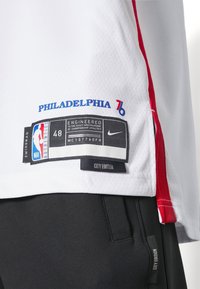 Maglia da basket bianca con accenti rossi, presenta la scritta "PHILADELPHIA 76" e etichette, realizzata in materiale mesh. Include etichetta della taglia e logo del marchio.