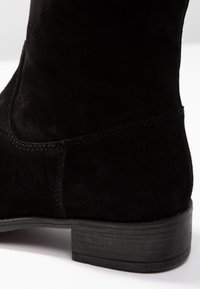 Stivaletti alla caviglia in suede nero con una texture liscia e morbida, punta arrotondata e tacco in legno impilato. Presenta un delicato dettaglio di cucitura lungo le cuciture.