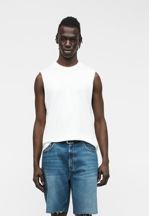 Rövid fonott hajjal rendelkező férfi ujjatlan fehér pólóban és kék farmer rövidnadrágban, lazán a teste mellett tartott kezekkel áll.