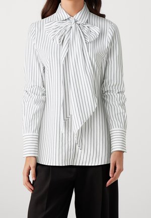White blouse with thin black vertical stripes, featuring a large bow tied at the collar, worn with black trousers.