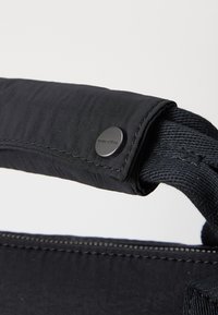 Poignée de sac en tissu noir avec une section rembourrée et un détail en métal circulaire. Texture de surface lisse, couture renforcée le long du bord.
