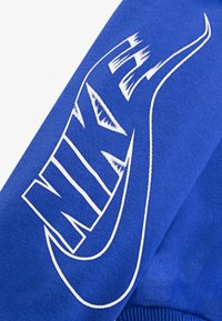 Mėlyna džemperis su baltu Nike tekstu ir logotipu drąsiu, moderniu šriftu. Medžiaga yra lygaus paviršiaus su raukšlėtu apvadu.