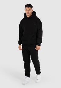 Zwarte hoodie en joggingbroek uit een zachte stof. De hoodie heeft een voorzak en een logo-detail. Witte sneakers maken de outfit compleet.