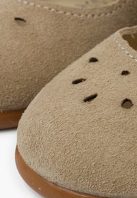 Jasnobrązowe zamszowe buty wsuwane mają zaokrąglony czubek z perforowanymi detalami oraz brązową gumową podeszwą. Widać ich miękką teksturę.