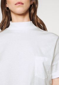 Bílý bavlněný tričko s vysokým límcem, krátkými rukávy a kapsou na levé straně hrudi. Jemná textura s jednoduchým, minimalistickým designem.