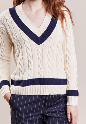 Vrouw draagt een crèmekleurige kabelgebreide trui met marineblauwe V-hals en strepen, gecombineerd met marineblauwe broek met fijne strepen tegen een beige achtergrond.